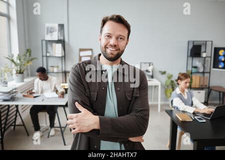 Porträt eines jungen bärtigen Geschäftsmannes, der mit gekreuzten Armen im Büro steht und die Kamera anlächelt Stockfoto