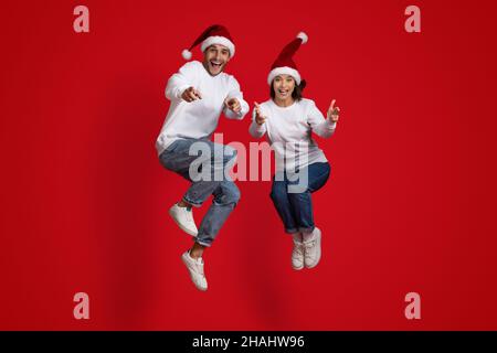 Überglücklich Mann Und Frau In Santa Hats Springen Und Zeigen Auf Kamera Stockfoto