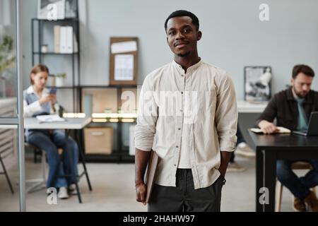 Porträt eines jungen afrikanischen Geschäftsmannes, der die Kamera anschaut, während er im Büro mit seinen Kollegen im Hintergrund steht Stockfoto