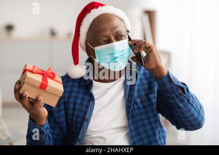 Senior African Male Holding Gift Talking Handy Trägt Maske Indoor Stockfoto