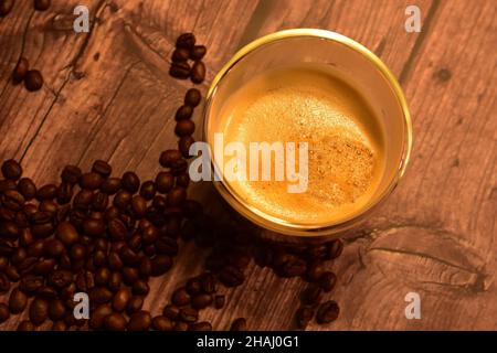 Draufsicht auf eine Glastasse mit köstlichem Kaffee mit Crema auf einem rustikalen Holztisch mit gerösteten Kaffeebohnen. Stockfoto