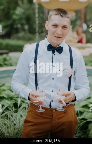 Hübscher Bräutigam mit zwei Gläsern Champagner. Ein gutaussehender, glücklicher Mann in elegantem Anzug, der zwei Gläser Champagner hält und seinen feiert Stockfoto