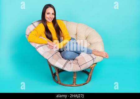 In voller Größe Foto von schönen zufrieden Dame sitzen weichen Stuhl toothy Lächeln Blick Kamera isoliert auf türkisfarbenem Hintergrund Stockfoto