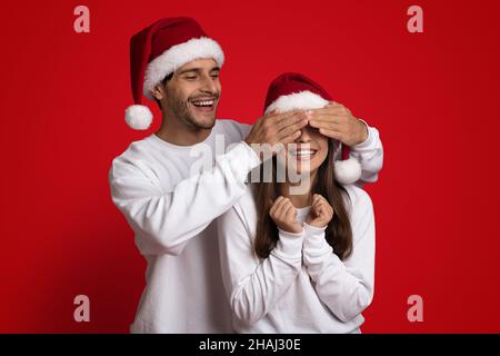 Raten Sie Mal, Wer. Verspielter Kerl in Weihnachtsmütze, der die Augen der Freundin mit den Händen bedeckt Stockfoto