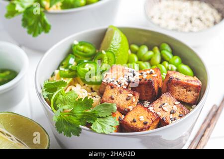 Vegane Nudeln Ramen Suppe mit marinierten Tofu, edamame Bohnen und Paprika in einer grauen Schüssel auf weißem Fliesenhintergrund, Nahaufnahme. Stockfoto