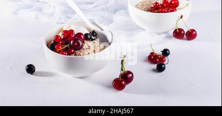 Roher Haferflocken mit Joghurt und Beeren aus Kirsche, Johannisbeere und Heidelbeere für ein gesundes Frühstück in einer weißen Keramikschale auf hellem Hintergrund. Selecti Stockfoto