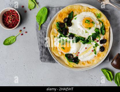 Türkische Eier Fladenbrot mit Joghurt, Käse, Oliven, Spinat und rotem Pfeffer auf Keramik-Vintage-Teller auf grauem altem Hintergrund. Draufsicht. Stockfoto