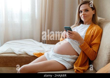 Porträt einer jungen 30 s Schwangeren, die in einem gemütlichen Zuhause auf dem Sofa sitzt. Kaukasische blonde Schwangere mit Bauch Verwenden Sie Smartphone-Handy für Stockfoto