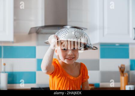 Glücklich Tochter Porträt spielt in der Küche. Kind Kind Mädchen in Mehl verschmutzt setzen Sieb Schüssel auf den Kopf wie Hut viel Spaß und lacht Spiel spielen. Glücklich Stockfoto