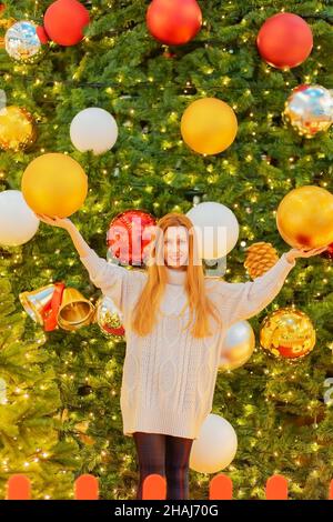 Lächelndes Mädchen hält riesige weihnachtskugeln, während sie in der Nähe des riesigen funkelnden weihnachtsbaums steht Stockfoto