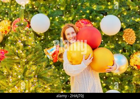 Fröhliches Mädchen hält große Weihnachtskugeln in der Nähe funkelnde Weihnachtsbäume. Festliche Atmosphäre. Stockfoto
