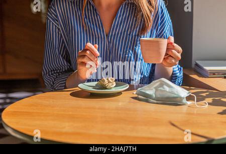 Frauen Hand-und Kaffeetasse und süße Dessert und medizinische Gesichtsmaske auf Holzschreibtisch im Kaffeehaus, Nahaufnahme. Stockfoto