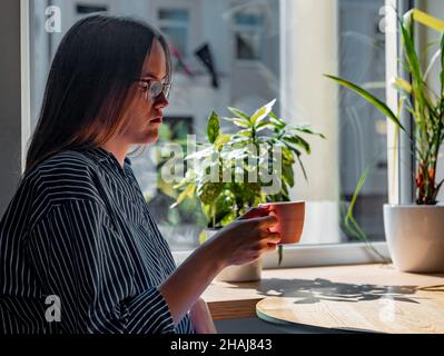 Frau in Gläsern sitzt am Holztisch im Café, hält Tasse und trinkt. Traurige, müde Frau in der Cafeteria denkend. Stockfoto