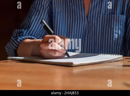 Frau Hand mit Stift schreiben in Notebook auf Holzschreibtisch mit Sonnenlicht, Nahaufnahme. Stockfoto