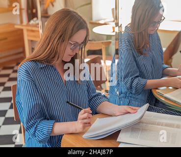 Junge Frau, die in einem Tagebuch schreibt, sitzt am Holzschreibtisch im Café mit Notizbüchern und Büchern. Stockfoto