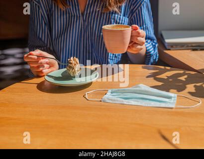 Frauen Hand-und Kaffeetasse und süße Dessert und medizinische Gesichtsmaske auf Holzschreibtisch im Kaffeehaus, Nahaufnahme. Stockfoto