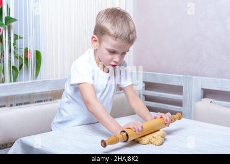 Das Kind rollt den Teig mit einem Nudelholz auf dem Tisch in der Küche aus. Der kleine Junge hilft der Mutter beim Kochen und macht selbstgemachte Gurkensaugen. Der Junge lernt Stockfoto