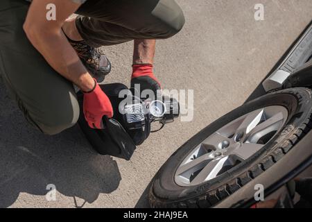 Draufsicht auf männliche Hände im Handschuh und tragbare Reifenpumpe zum Aufblasen des Autorads. Luftkompressor für Reifenfüller mit Manometer. Stockfoto