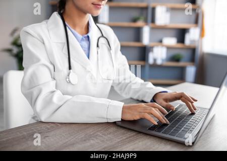 Eine junge hindu-Ärztin in weißem Mantel mit Stethoskop, die am Computer im Büroinneren tippt Stockfoto