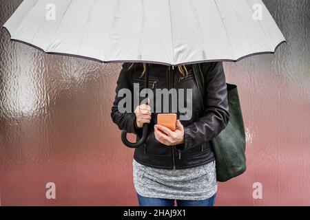 Frau mit Regenschirm, Smartphone haltend und im Regen in der Stadt stehend. Mit dem Mobiltelefon Stockfoto