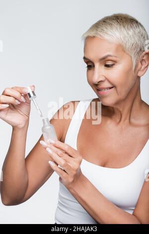 Glückliche ältere Frau, die Serum mit Pipette hält, die auf Grau isoliert ist Stockfoto