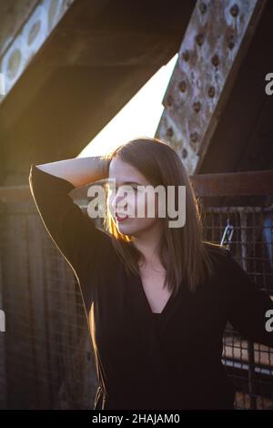 Junge schöne Frau, die an der Brücke steht und den Sonnenuntergang in der Stadt genießt. Modische attraktive Brünette Frau im Freien Stockfoto