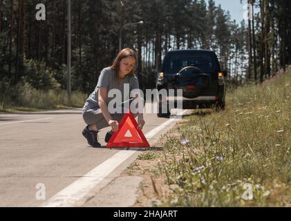 Junge Frau, die ein Warnschild mit einem roten Dreieck auf der Straße in der Nähe eines kaputten Autos am Straßenrand angebracht hat. Stockfoto