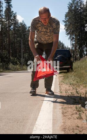 Mann, der am Straßenrand ein Warnschild mit Dreieck auf der Straße in der Nähe eines kaputten Autos angebracht hat. Stockfoto