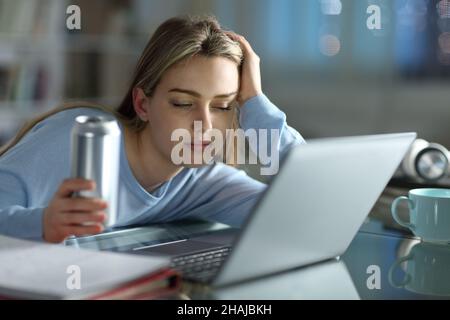 Schlafende Schülerin, die versucht, mit einem Laptop zu lernen, der nachts zu Hause ein Energiegetränk hält Stockfoto