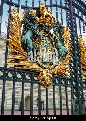 Königliches Wappen an den Eingangstoren des Buckingham Palace mit traditioneller Wache in roter Tunika und busby im Hintergrund, London, England Stockfoto