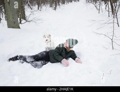 Teenager Mädchen 16-17 Jahre alt liegt im Schnee mit weißem Hund. Spaziergang mit Haustier in der kalten Wintersaison. Spaß mit geliebtem Hund. Ein aktiver Lebensstil, Freude aller Zeiten Stockfoto