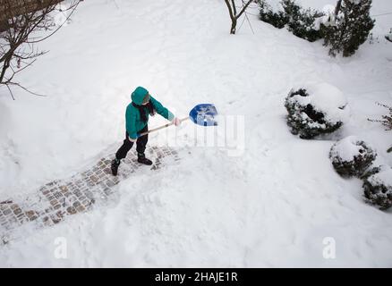 Nicht erkennbare junge Frau in einer Kapuze, die den Bürgersteig, Straße von Schnee nach einem starken Schneesturm, mit einer Schneeschaufel. Wintersaison, schlechtes Wetter Stockfoto