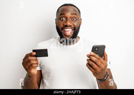 Glückselig überraschte der junge Afroamerikaner, der Debitkarte und Smartphone hält und mit offenem Mund isoliert auf die Kamera schaut. Glücklicher schwarzer Mann im lässigen T-Shirt erhalten Cashback aus Online-Einkäufen Stockfoto