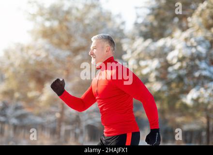 Sportlicher älterer Mann in Sportkleidung, der im Winterpark läuft, Seitenansicht. Saisonales Sport- und Temperierungskonzept Stockfoto
