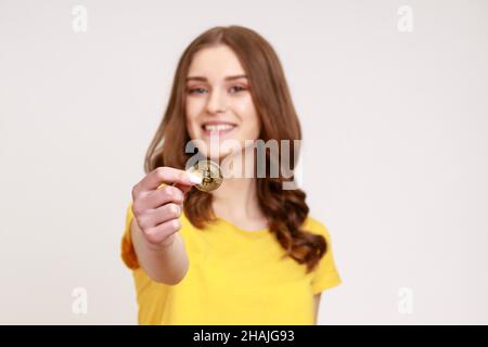 Kryptowährung BTC Coin. Reiche zufriedene glückliche braune haarige junge Frau in gelbem T-Shirt Blick auf die Kamera, hält goldene Bitcoin, elektronisches Geld. Innenaufnahme des Studios isoliert auf grauem Hintergrund. Stockfoto