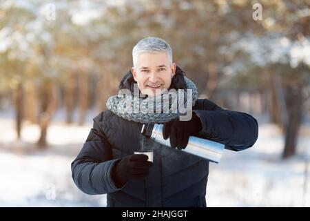 Lächelnder älterer Mann in warmen Winterkleidung, der im verschneiten Park Tee aus Thermoskanne gießt Stockfoto