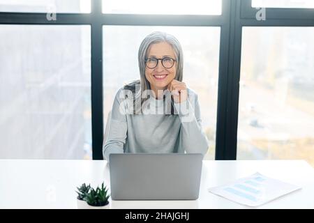 Porträt einer charmanten, eleganten und reifen Geschäftsfrau in einer Brille, die lächelnd auf die Kamera schaut, die auf dem modernen Schreibtisch vor einem Laptop sitzt, genießt eine freiberufliche Mitarbeiterin mittleren Alters die Arbeit von zu Hause aus Stockfoto