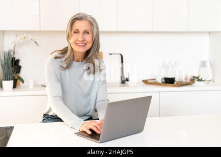 Fröhliche, reife, grauhaarige Frau verbringt ihre Freizeit online, lächelnde Frau sitzt mit einem Laptop in der Küche, schaut sich Filme an, scrollt in den Nachrichten in den sozialen Medien, schaut auf die Kamera und lächelt Stockfoto