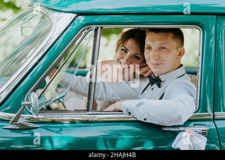 Wunderschöne stilvolle blonde Braut posiert im Retro-grünen Auto mit Bräutigam. Braut und Bräutigam sitzen im Retro-Auto. Hochzeit, Braut und Bräutigam Stockfoto