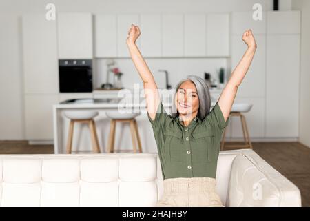Grauhaarige Frau, die die Arme streckt, sitzt zu Hause auf dem Sofa, eine fröhlich lächelnde asiatische Dame bereitet sich auf die Stretching-Übung vor und macht eine Pause Stockfoto