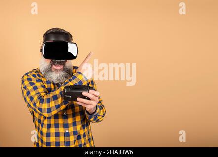 Gamer in VR-Brille auf gelbem Hintergrund hält ein Gerät und zeigt seine Finger auf einen Ort für Text, einen Mann mit drahtlosen Gadgets für die virtuelle Realität Stockfoto
