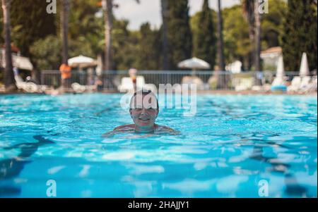 Junge Frau, die im leeren Pool des tropischen Resorts auf die Kamera zuschwimmend, lächelt Stockfoto