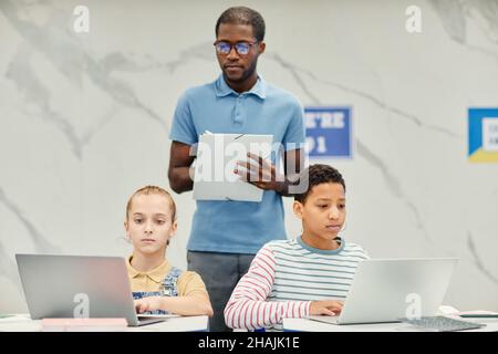 Porträt von zwei Mädchen im Teenageralter, die im modernen Schulklassenzimmer Laptops benutzen, mit afroamerikanischem Lehrer, der ihnen bei der Arbeit zusieht und Raum kopiert Stockfoto