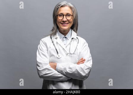Lächelnd grauhaarige Ärztin für Kinder, Physiotherapeuten, die einen weißen medizinischen Kittel mit Stethoskop auf den Schultern trägt, steht mit gekreuzten Armen isoliert auf grauem, kopiertem Raum. Gesundheitswesen und Medizin Stockfoto