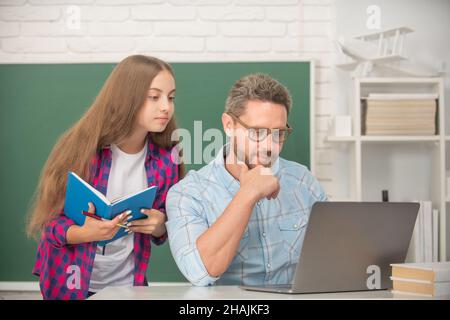 Aufmerksames Kind und Vater sitzen im Klassenzimmer mit Copybook und Computer an der Tafel, Schularbeiten Stockfoto