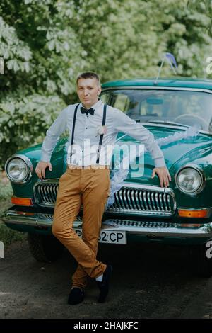 Retro-Fahrer im Oldtimer auf dem Outdoor-Hintergrund. Schöner Bräutigam im Auto. Selbstbewusster, wohlhabender junger Mann in der Nähe von Oldtimer. Elegant Stockfoto