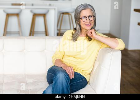 Zauberhafte, sorglose grauhaarige Frau mittleren Alters sitzt auf der Couch und entspannt sich. Porträt einer charmanten reifen Frau, die zu Hause träumt Stockfoto
