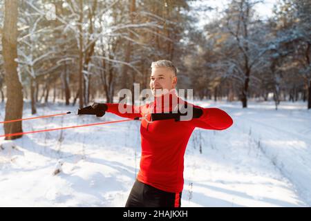 Funktionelles Workout. Starker, reifer Sportler, der an kalten, verschneiten Tagen mit Bodyweight Resistance Straps im Park trainiert Stockfoto