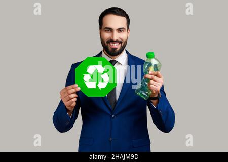 Lächelnder bärtiger Mann, der die Hand mit einer Plastikflasche ausstreckte, auf der ein Recycling-Schild zu sehen war, Abfall sortierte und die Natur schützte, und einen offiziellen Anzug trug. Innenaufnahme des Studios isoliert auf grauem Hintergrund. Stockfoto
