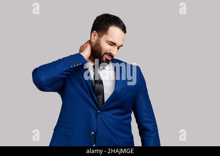 Bärtiger Geschäftsmann, der den Hals berührt und Schmerzen und Taubheit empfindet, sich Sorgen über Muskelverspannungen, Osteochondrose macht und einen offiziellen Anzug trägt. Innenaufnahme des Studios isoliert auf grauem Hintergrund. Stockfoto